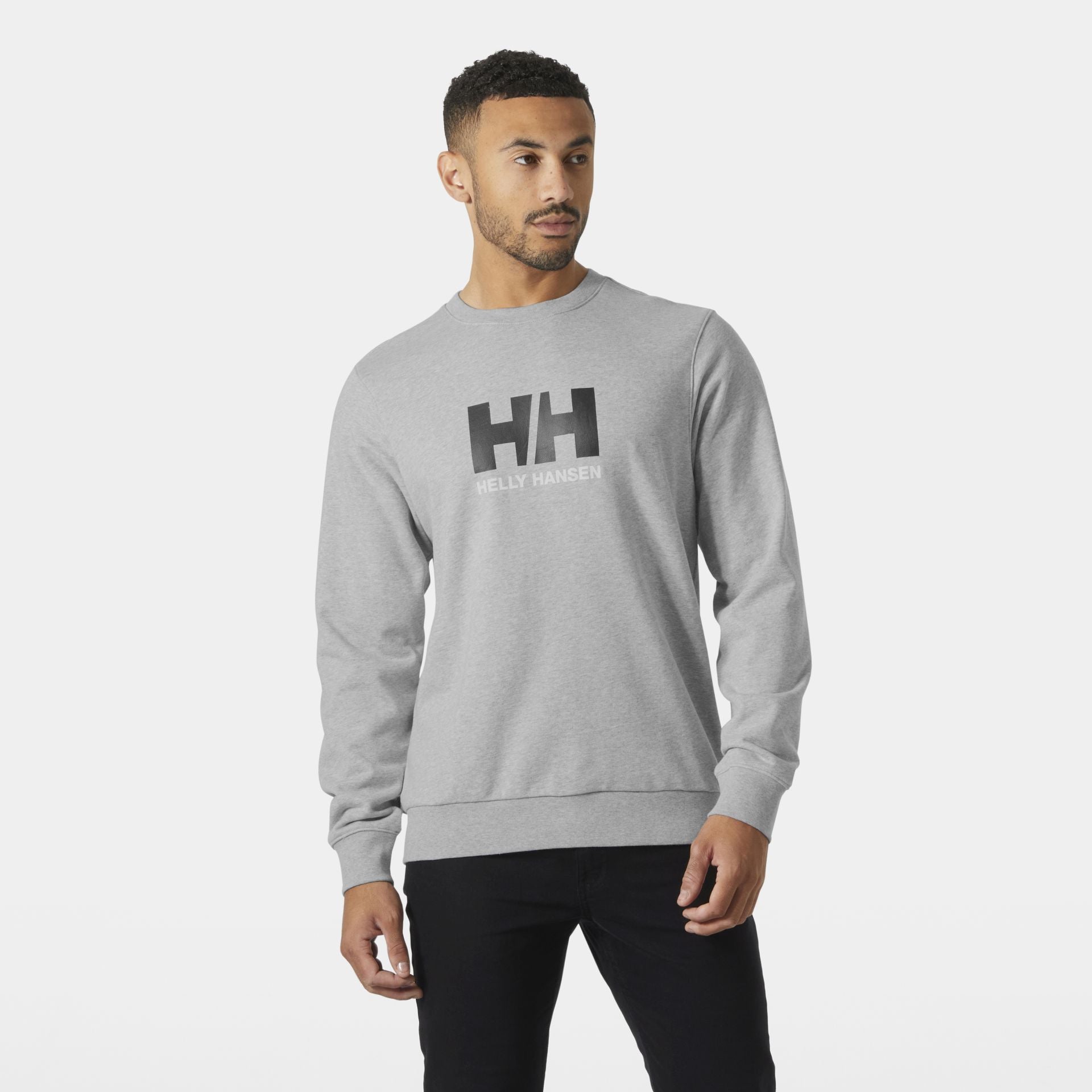 Mann trägt ein graues Helly Hansen Sweatshirt mit schwarzem HH-Logo auf der Brust, steht vor neutralem Hintergrund und blickt leicht zur Seite.