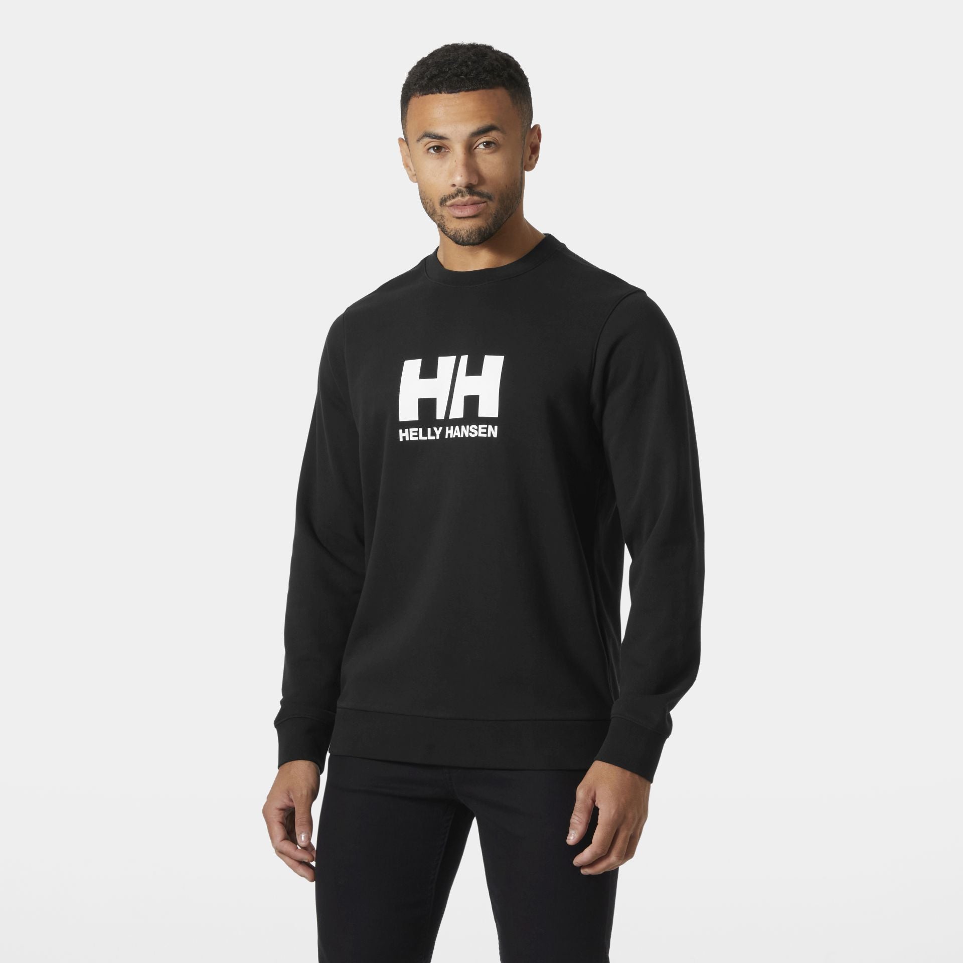 Mann trägt ein schwarzes Helly Hansen Sweatshirt mit weißem HH-Logo auf der Brust, steht vor neutralem Hintergrund und blickt leicht zur Seite