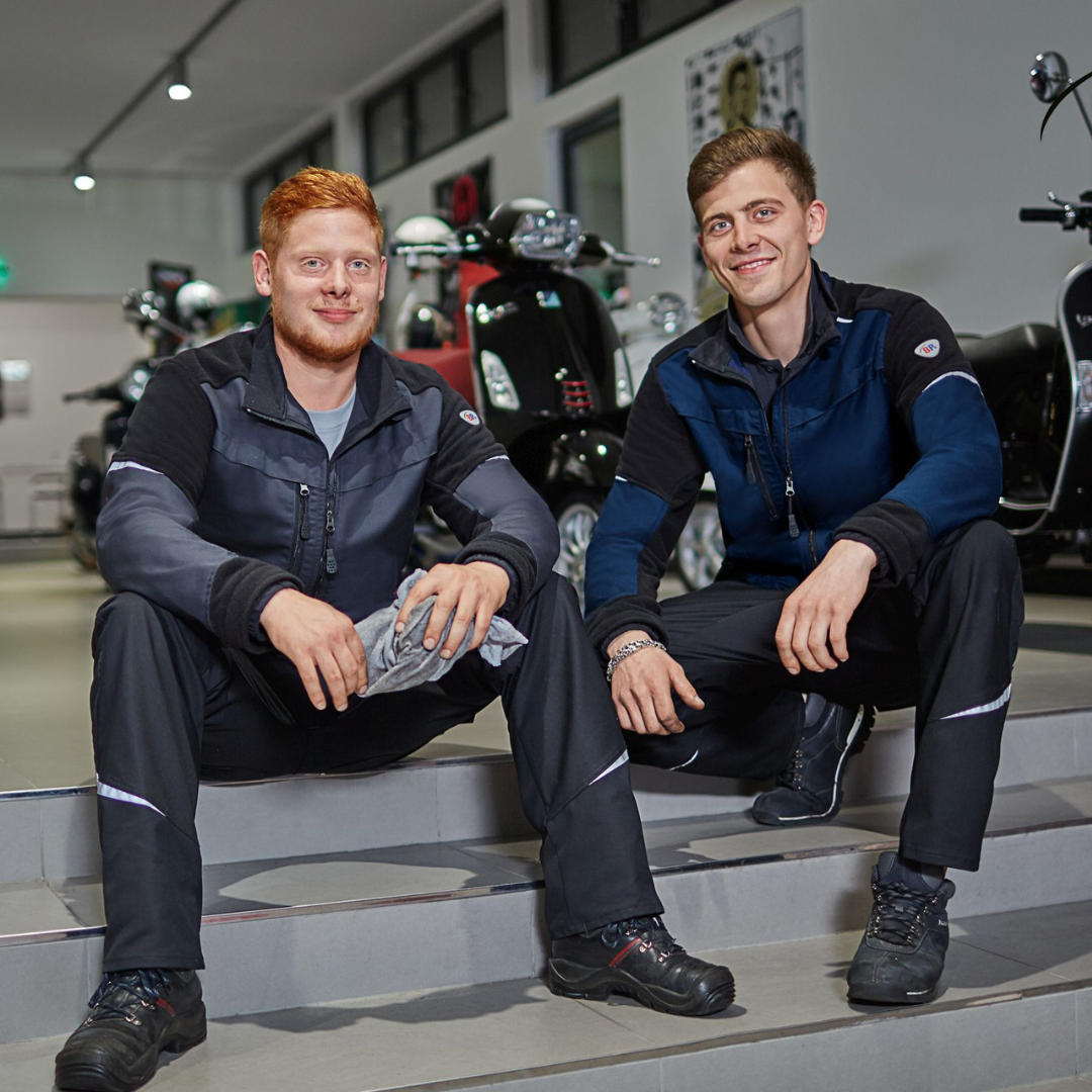 Zwei junge Männer in moderner Arbeitskleidung mit Softshelljacke und Sicherheitsschuhen sitzen entspannt auf Treppenstufen in einem Verkaufsraum für Motorroller – funktionale Berufskleidung für Handwerk und Werkstattumgebung.